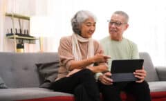 senior couple sitting on sofa using tablet while video call online with family in living room at home.
