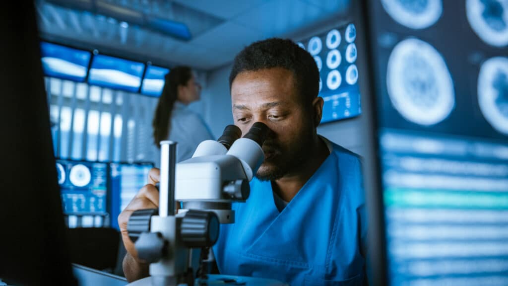 Medical Research Scientist Looking under the Microscope in the Laboratory. Neurologist Solving Puzzles of the Mind and Brain. In the Laboratory with Multiple Screens Showing MRI / CT Brain Scan